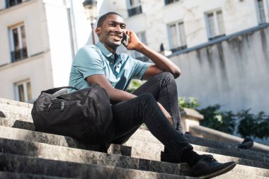 Tam vücut portre mutlu genç siyah adam merdivenlerde oturuyor cep telefonuyla konuşuyor. 