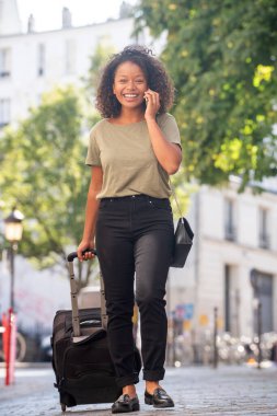 Tüm vücut portresi mutlu genç bir kadın şehirde çanta ve cep telefonuyla yürüyor.