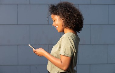 Gri duvarda cep telefonuna bakan gülümseyen genç bir kadın.