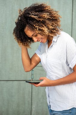 Genç Afrikalı Amerikalı kadın gülümsüyor ve yeşil duvarda cep telefonuna bakıyor.