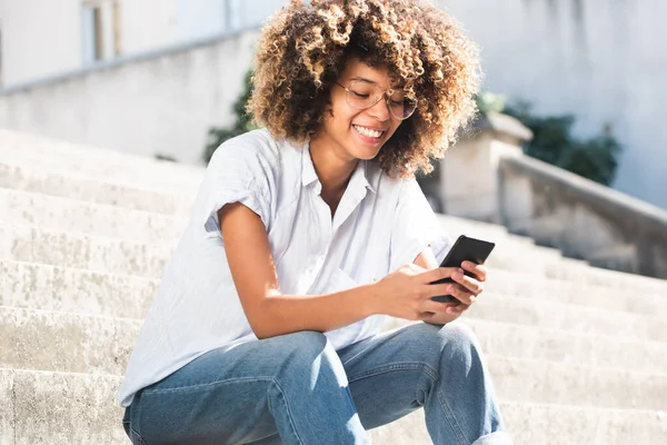 ポートレート幸せな若いです黒女性とともに眼鏡をかけて座っています外で電話を見て — ストック写真