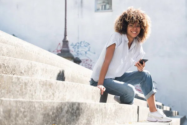Retrato Sonriente Joven Afroamericana Con Anteojos Sentados Afuera Sosteniendo Teléfono — Foto de Stock
