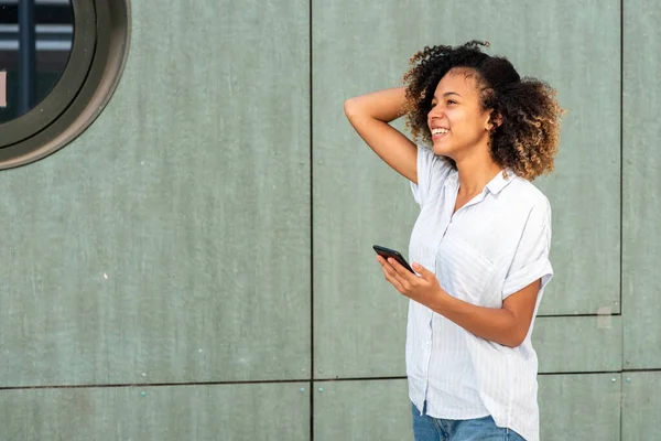 サイドポートレート幸せな若いアフリカ系アメリカ人女性保持携帯電話によって緑の壁 — ストック写真