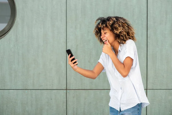 Portrait laughing young African American woman looking at mobile phone by green wall
