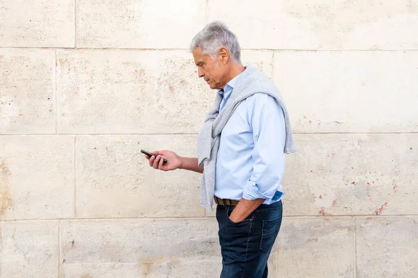 profile portrait of older man looking at mobile phone while walking by white wall