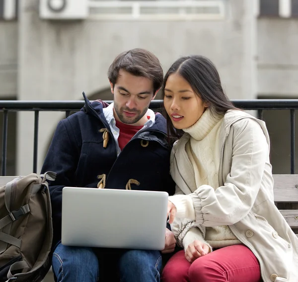 Pareja joven sentada fuera en el banco mirando la pantalla del ordenador portátil — Foto de Stock