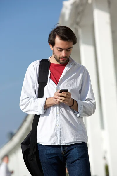 Young man with bag walking and sending text message outdoors — Stock Photo, Image