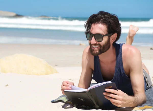 Bonito jovem sorrindo e lendo livro na praia — Fotografia de Stock