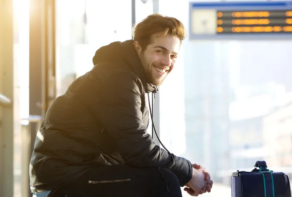 Šťastný mladý muž sedí s taškou na vlakovém nádraží — Stock fotografie