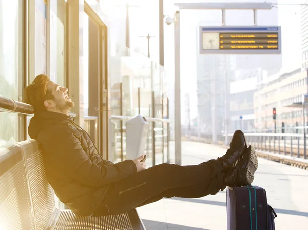 Glücklicher junger Mann sitzt mit Reisetasche am Bahnhof — Stockfoto