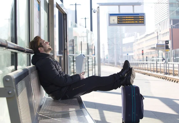 Usmívající se mladý muž sedí s taškou, čekání na vlak — Stock fotografie