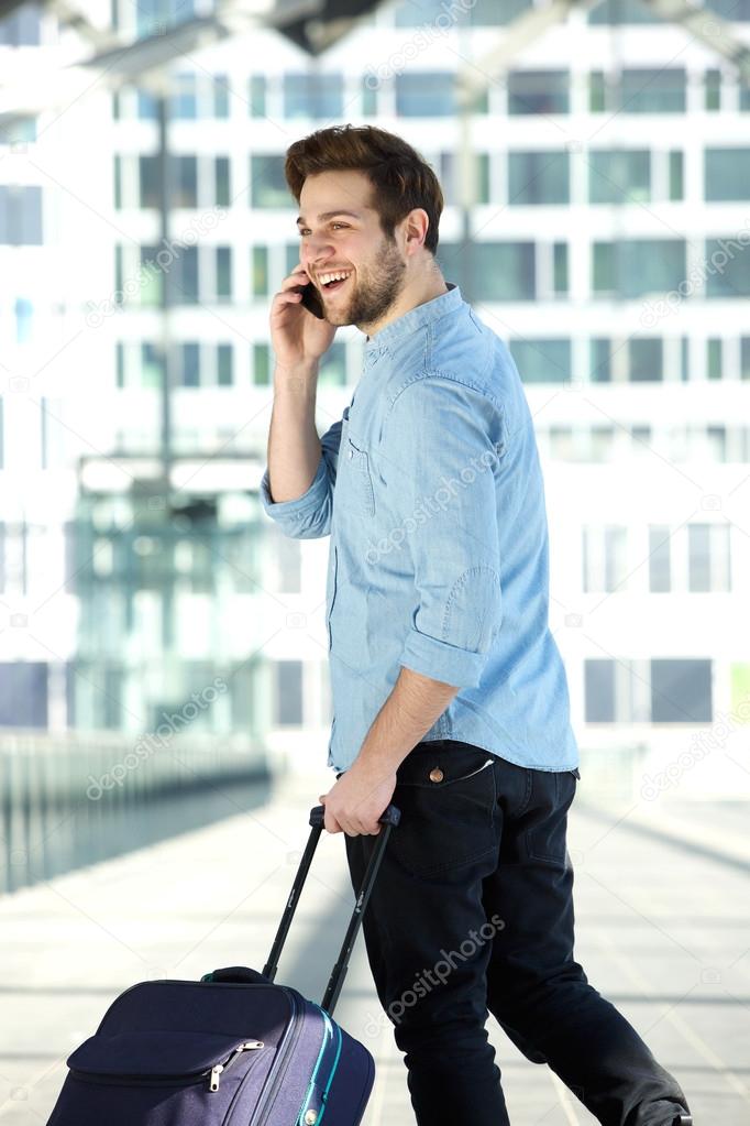 Happy young man walking at airport with bag and mobile phone