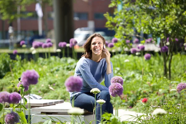 Uśmiechnięta kobieta siedzi poza w parku i przy użyciu telefonu komórkowego — Zdjęcie stockowe