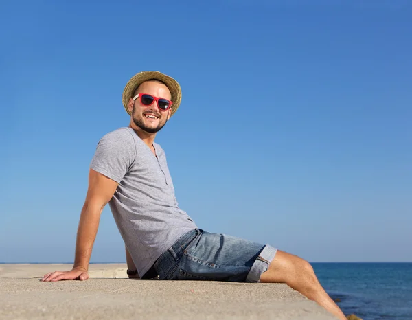 Happy young man sitting by the sea in summer with hat — Stock Photo, Image