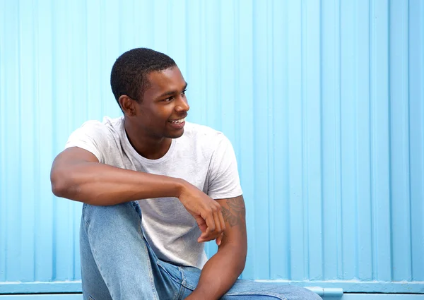 Sonriente joven sentado contra la pared azul mirando hacia otro lado —  Fotos de Stock