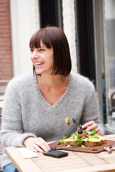 Улыбающаяся женщина, сидящая за столом, ест пищу — стоковое фото
