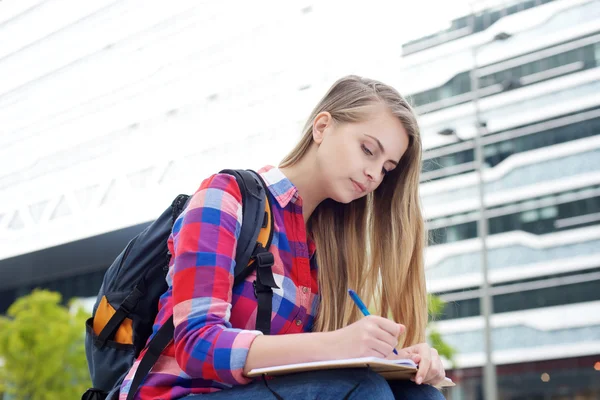 Female student sitting outside with bag and writing in book — Stock Photo, Image