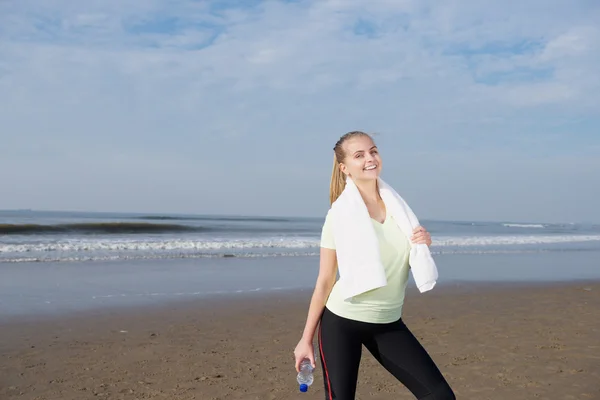Улыбающаяся спортивная женщина, стоящая с бутылкой воды на пляже — стоковое фото