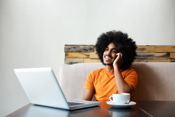 Sonriente chico negro sentado en la cafetería con ordenador portátil y teléfono celular — Foto de Stock