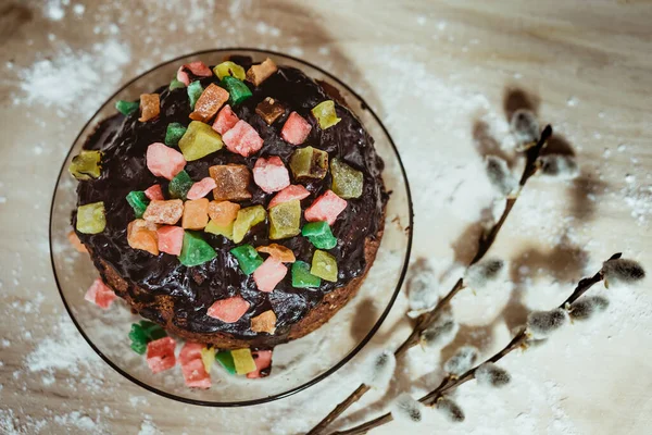 Çikolata kremalı kek, şekerli meyvelerle süslenmiş. Un serpiştirilmiş ahşap bir standın üzerinde duruyor.