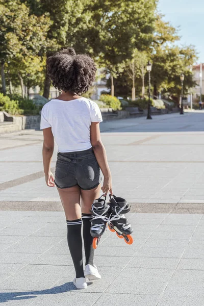 Afro saçlı genç Afrikalı bir kadın, güneşli bir günde paten antrenmanına gitmek ve spor yapmak için elinde patenlerle parkta yürüyor.