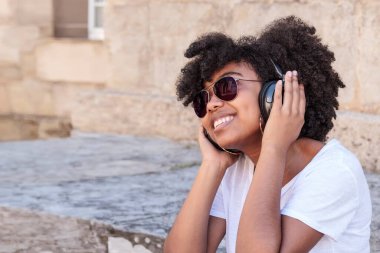 Siyah saçlı, gülümseyen bir kız sıcak bir yaz gününde kulaklıkları ve güneş gözlükleriyle müzik dinliyor.