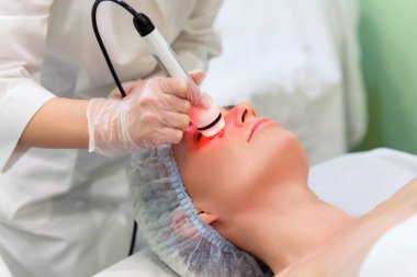 Kaplıca salonundaki genç bir kadın yüzünü gerdiriyor. Güzellik uzmanı RF-kaldırma aparatını yüzünün üzerinde tutuyor..