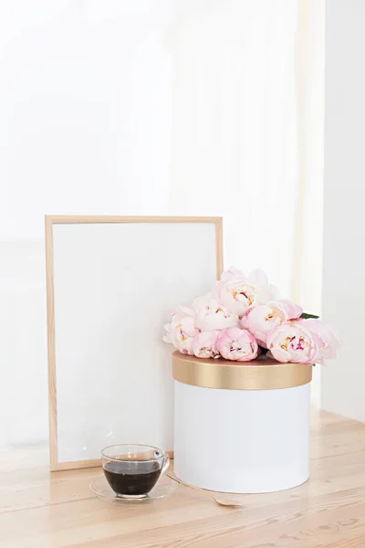 Vertikaler Rahmen und Geschenkbox-Attrappe auf einem Holztisch in der Küche. Glaskanne mit einem Strauß rosa Pfingstrosen und einer Tasse schwarzen Kaffees. Innenraum im skandinavischen Stil. — Stockfoto