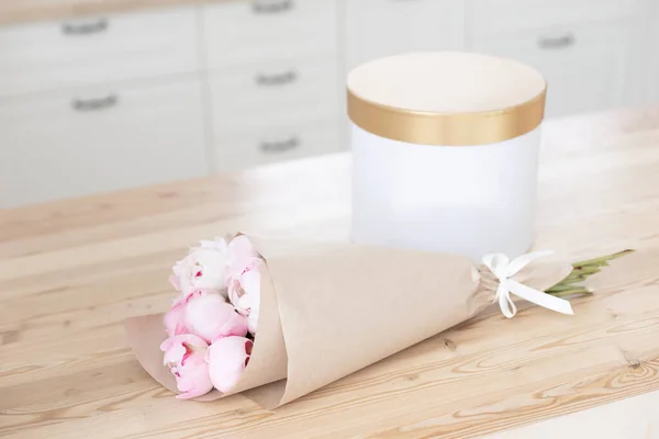 Zlatá dárková krabice na dřevěném stole v kuchyni. Skleněná váza s kyticí růžových pivoňek. Interiér skandinávského stylu. — Stock fotografie