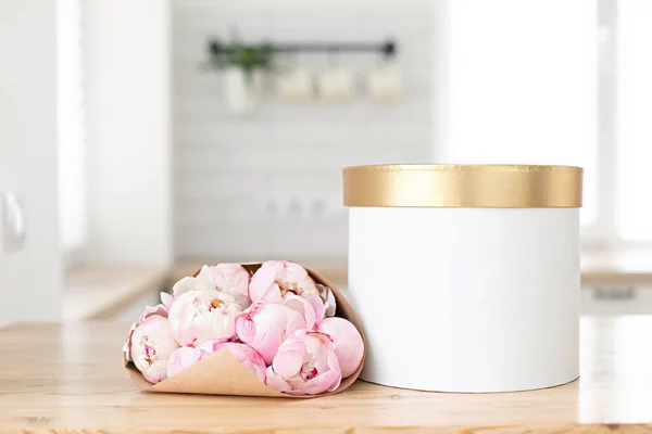 Χρυσό κουτί δώρου mockup σε ένα ξύλινο τραπέζι στην κουζίνα. Γυάλινο βάζο με μπουκέτο από ροζ παιώνιες. Σκανδιναβικό εσωτερικό στυλ. — Φωτογραφία Αρχείου