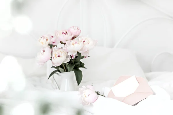 Bouquet de pivoines roses et enveloppe de salutations sur du linge de lit blanc. Intérieur moderne dans la chambre. Mariage et style festif. — Photo