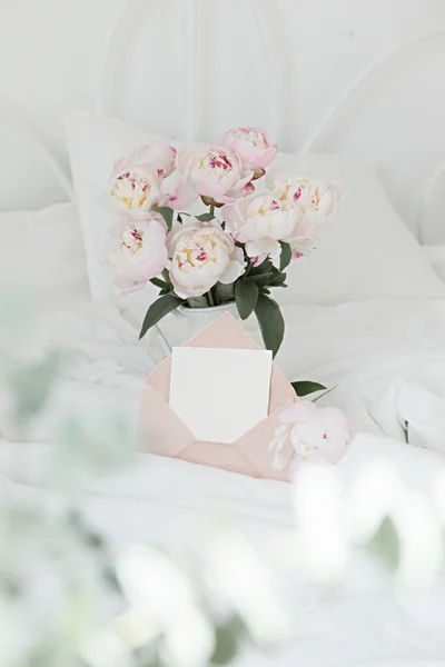 Bouquet de pivoines roses et enveloppe de salutations sur du linge de lit blanc. Intérieur moderne dans la chambre. Mariage et style festif. — Photo