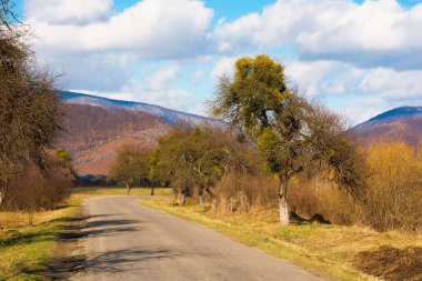 İlkbaharın başlarında kırsal alandan geçen eski bir yol. Yol boyunca yapraksız ağaçlar. Uzakta karla kaplı bir dağ. Gökyüzünde bulutlar olan güneşli bir hava