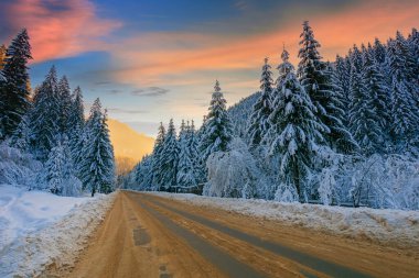 Kışın dağ manzarası boyunca yol. Karla kaplı ladin ormanı. Akşam ışığında parlayan bulutlarla dolu dramatik bir gökyüzü