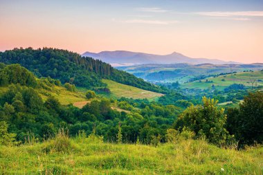 Şafak vakti dağlık kırsal alan. Ormanlar, tepeler ve sabah güneşinde çayırlar olan güzel bir manzara. Uzakta tepe noktası olan bir tepe. Uzak vadideki köy