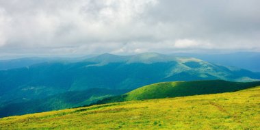 Yazın bulutlu bir günde dağ manzarası. Çimenler yamaçtaki çayırları kaplamış. Uzaktaki Karpatya havza sırtı