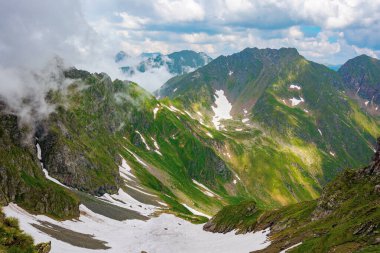 İbnelerin yaz mevsiminde dağ manzarası. Vadinin tepelerinde bulutlar ve karla kaplı harika doğa manzarası. Çimlerdeki ve kayalardaki tepeler. Romanya konseptinin karpatlarını keşfetmek