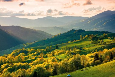 Gün batımında dağlarda kırsal alan. Baharda uzak vadide dramatik bir hava var. Tepedeki yeşil tarlalar ve ağaçlar. Gökyüzünde bulutlar olan güzel doğa manzarası