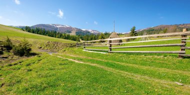 Baharda dağlık kırsal alan. Yuvarlanan tepelerdeki ahşap çitlerin arkasındaki çimenlik arazide saman yığını. Uzakta kar tepeli sırt. Güneşli bir günde güzel bir kır manzarası.