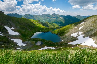 İbne Dağları 'nın Capra Gölü. Güneşli bir günde harika bir yaz manzarası. Romanya 'nın popüler seyahat beldesi. Yamaçlarda kar ve çimenler. Gökyüzünde kabarık bulutlar