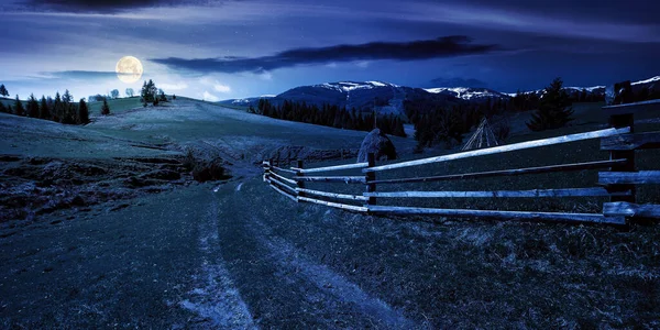 mountainous rural landscape at night in spring. path through grassy field. wooden fence on rolling hills. snow capped ridge in the distance. wonderful countryside scenery in full moon light