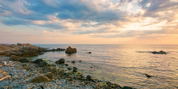 日の出の海で夏休みの風景 穏やかな水は石や岩で小石のビーチを洗います 地平線の上の劇的な雲が空を照らす — ストック写真