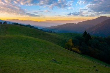 Şafak vakti dağlık bir kırda. Tepelerdeki ağaçlar ve çimenli çayırlar. Parlak gökyüzünün altında, akşam ışığında bulutlarla tepeler.