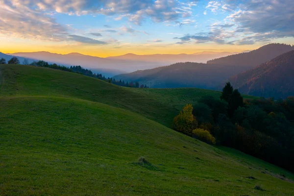 Şafak vakti dağlık bir kırda. Tepelerdeki ağaçlar ve çimenli çayırlar. Parlak gökyüzünün altında, akşam ışığında bulutlarla tepeler.