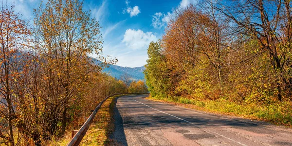 old country road in mountains. trees in colorful foliage in morning light along the serpentine curve. sunny weather with clouds on the sky above the distant range