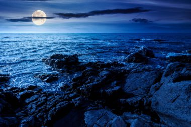 weekend on a rocky beach at the black sea under open dark sky at night. beautiful coastline in summer. seascape scenery with clouds above horizon in full moon light. mysterious weather