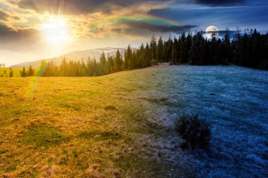 Bahar ekinoksunda ladin ormanları olan dağ manzarası. Gündüz ve gece zaman kavramını değiştirir. Tepedeki yeşil çayırda güneş ve ay var. Gökyüzünün altında bulutlarla kaplı uzak bir menzil. Güzel manzara.
