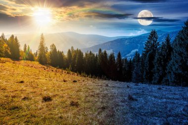 Sonbahar ekinoksunda çayırın arkasındaki kozalaklı ormanın manzarası. Gündüz ve gece zaman kavramını değiştirir. Mavi gökyüzü ve güneş ve ay ile kaplı dağ sırtı. Alp ormanları
