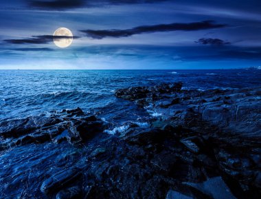 Kayalık deniz kıyısının gece manzarası. Ufukta güzel bir dolunay görünüyor. Yaz kıyı şeridi manzarası. Gece yarısı birleşik sahne. Sihirli deniz manzaralı tatiller. Karanlıkta dalgalar ve kayalar