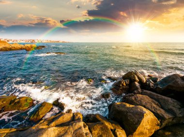 Kayalık deniz sahilinde gün batımı. Yazın güzel bir kıyı manzarası. Güneş ufukta ve bulutlu gökyüzünde. Gökkuşağı altında kaya oluşumlarıyla dramatik bir manzara. Hafta sonu için harika deniz manzarası